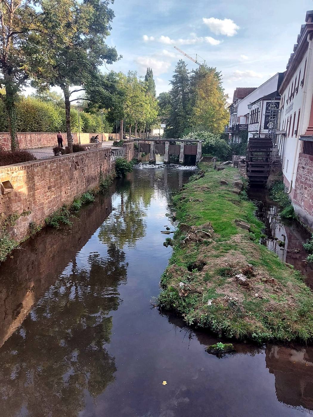 Die Mümling in Erbach im Odenwald zwischen Sandsteinmauern, mit hölzernem Wehr, altem Wasserrad und Uferweg mit Bänken